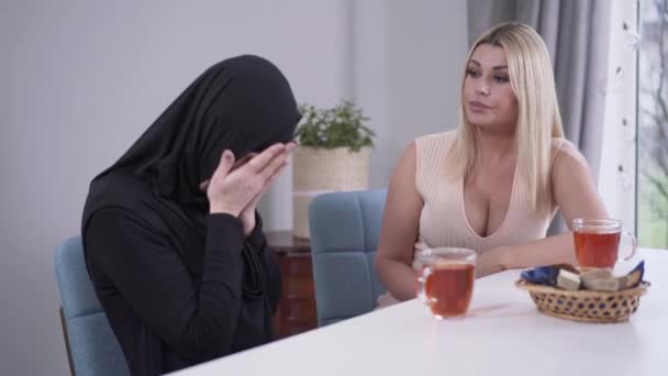 Ung Muselman kvinna klaga till Kaukasisk vän inomhus. Orolig dam i traditionell hijab talar med modern flicka i uppriktig klänning. Vänskap, frustration, stöd, tolerans. — Stockvideo