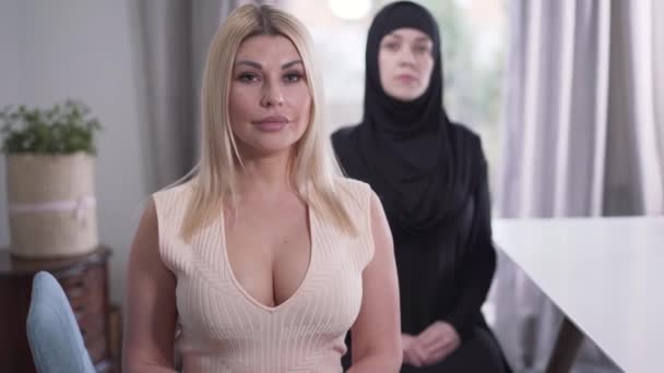 Portrait de femme caucasienne moderne confiante regardant la caméra. Femme musulmane floue dans le hijab traditionnel assis à l'arrière-plan. Différence des cultures, diversité culturelle, tolérance . — Video