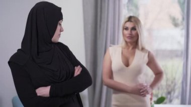 Muhafazakar Müslüman kadın, samimi giyinmiş ve arkasını dönmüş modern Kafkas kızına bakıyor. Türbanlı mütevazı bir kadın portresi ve kültürel çeşitliliği tasvir eden samimi elbiseli bulanık bir bayan..