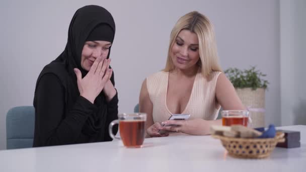 Застенчивая мусульманка и уверенная кавказка смотрит на экран смартфона и улыбается. Мультикультурные подруги отдыхают в помещении и пользуются социальными сетями. Дружба, разнообразие, терпимость . — стоковое видео