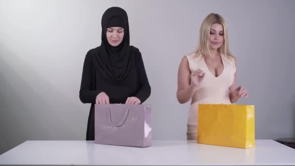 Two happy women coming with shopping bags and taking out new dresses. Modern Caucasian woman holding dress with sparkles as conservative Muslim woman enjoying new black traditional clothes. — Stock Video