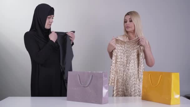 Porträt muslimischer und kaukasischer Frauen, die neue Kleider bewundern. Ost-Dame in schwarzem Outfit und blondes Mädchen in offenem Kleid schauen einander mit Sarkasmus an. Mode, kultureller Unterschied. — Stockvideo