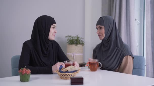 Dvě muslimky v hijabs sedí u stolu s čajem a mluví. Pozitivní mladé dámy drbající vevnitř. Východní kultura, komunikace, přátelství. — Stock video