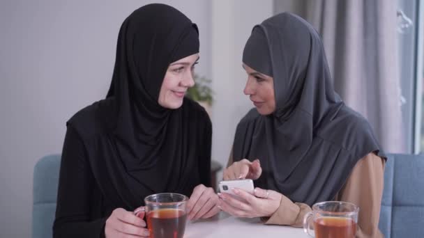 Mujer musulmana de aspecto moderno mostrando la pantalla del teléfono inteligente a un modesto amigo en hijab. Dos mujeres reaccionan emocionalmente en las redes sociales. Tecnologías modernas, comunicación, estilo de vida . — Vídeos de Stock