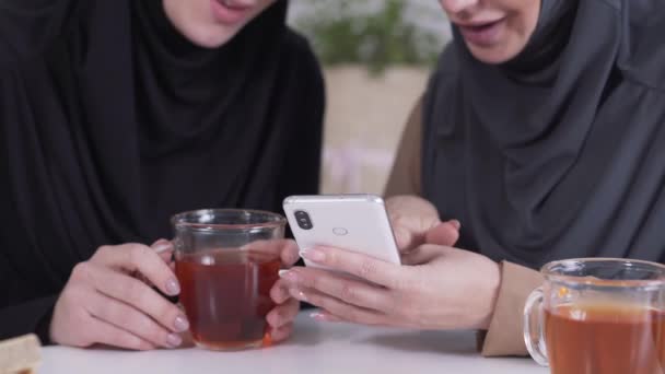 Zbliżenie dwóch nierozpoznawalnych muzułmanek korzystających ze smartfona, mówiących i uśmiechających się. Współistnienie kultury tradycyjnej i nowoczesnego społeczeństwa. Styl życia, social media, Internet. — Wideo stockowe