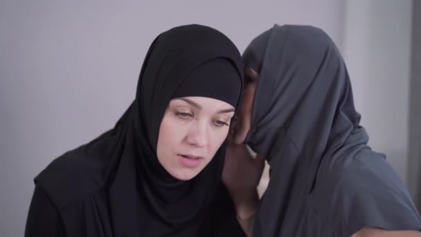 Portret van een jonge moslimvrouw in hijab die luistert naar een vriend die op haar oor fluistert. Verrassende vrouw die haar mond bedekt met de hand. Twee vriendinnen roddelen. Lifestyle, traditionele cultuur. — Stockvideo