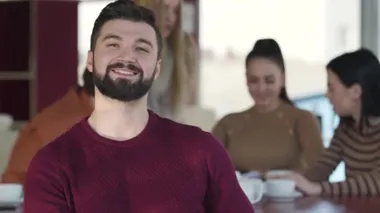 Portrait of confident handsome Caucasian man with black hair and beard looking at camera and smiling. Young positive man posing as friends chatting at the background. Lifestyle, leisure, happiness.
