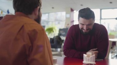 Portrait of young happy Caucasian man talking with best friend in cafe. Close-up of smiling young man with black hair and beard meeting mate in restaurant. Leisure, male friendship, lifestyle.
