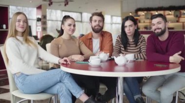 Portrait of five young Caucasian people sitting in cafe, looking at camera and smiling. Friendly fellows posing in restaurant. Happiness, leisure, lifestyle, friendship.