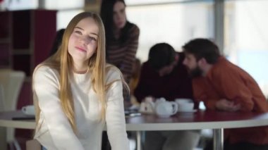 Beautiful young blond woman making victory gesture and smiling at camera. Cheerful female student posing at the background of groupmates discussing common project. Lifestyle, beauty.