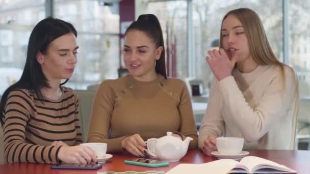 Retrato de tres jóvenes hermosas mujeres caucásicas sentadas a la mesa en la cafetería y hablando. Encantadoras damas descansando en el restaurante los fines de semana. Felicidad, estilo de vida, amistad femenina . — Vídeos de Stock