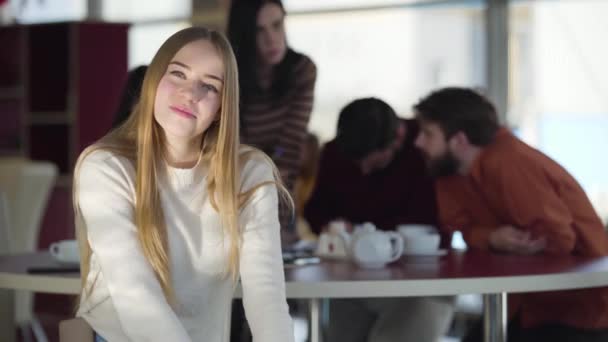 Beautiful young blond woman making victory gesture and smiling at camera. Cheerful female student posing at the background of groupmates discussing common project. Lifestyle, beauty. — Stok video