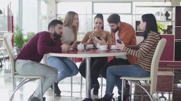 Pět mladých bělochů se dívá na obrazovku smartphonu a mluví. Přátelé používají gadgets jako odpočinek v kavárně za slunečného dne. Životní styl, moderní společnost, komunikace, digitální závislost. — Stock video