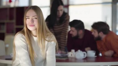 Portrait of charming blond Caucasian woman looking at camera and smiling. Group of friends chatting at the background. Beauty, happiness, lifestyle.