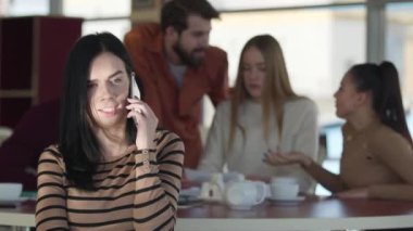 Young serious Caucasian woman talking on the phone while her friends chatting at the background in cafe. Confident adult girl resting with fellows in restaurant. Joy, lifestyle, communication.