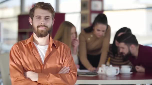 Portrait of young redhead bearded Caucasian man looking at camera and smiling as his friends studying at the background. Happy adult university student posing indoors. Lifestyle, joy, happiness. — Stok video
