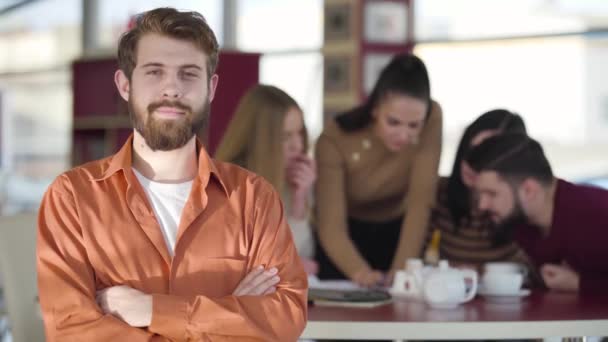 Retrato de la joven pelirroja barbuda hombre caucásico mirando a la cámara y sonriendo como sus amigos estudiando en el fondo. Feliz estudiante universitario adulto posando en el interior. Estilo de vida, alegría, felicidad . — Vídeo de stock