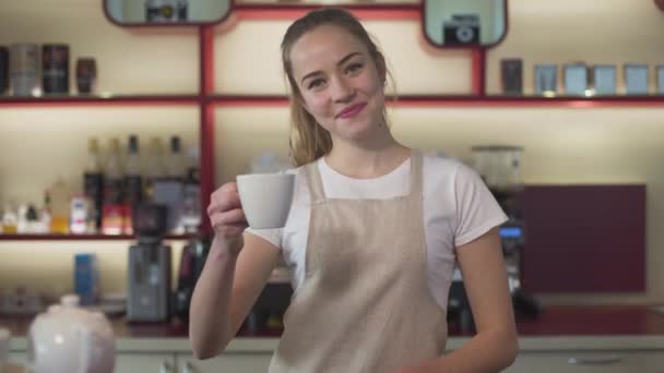 Zâmbind o tânără caucaziană care arată o ceașcă de cafea la cameră și zâmbind. Barista profesionistă care pozează la locul de muncă. Bucurie, satisfacție, ocupație, stil de viață . — Videoclip de stoc