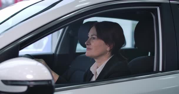 Vista lateral da mulher caucasiana adulta sentada no assento do motorista, olhando para a câmera e sorrindo. Retrato de proprietário feminino confiante de carro novo. Indústria automobilística, concessionária. Cinema 4k ProRes HQ . — Vídeo de Stock