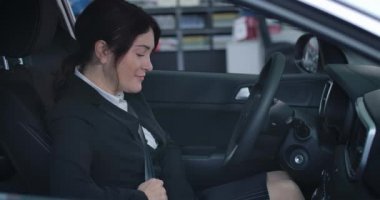 Side view of satisfied Caucasian brunette woman fastening seat belt and starting engine in new vehicle. Portrait of smiling businesswoman in automobile. Car dealership, lifestyle. Cinema 4k ProRes HQ.