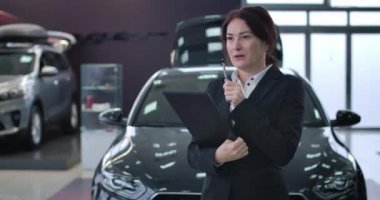 Confident Caucasian saleswoman looking around in car dealership and writing down in documents. Portrait of professional female trader in showroom at the background of vehicles. Cinema 4k ProRes HQ.