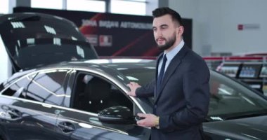 Başarılı Kafkas esmer, kahverengi gözlü adam yeni arabasının yanında duruyor ve gülümsüyor. Genç yakışıklı iş adamı galeride araç satın alıyor. Sinema 4k Prores Merkezi.