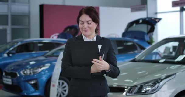 Retrato de morena adulta mujer caucásica con ojos marrones posando en concesionario de automóviles. Distribuidor sonriente en traje sosteniendo documentos y mirando a la cámara. Sede del cine 4k ProRes . — Vídeo de stock