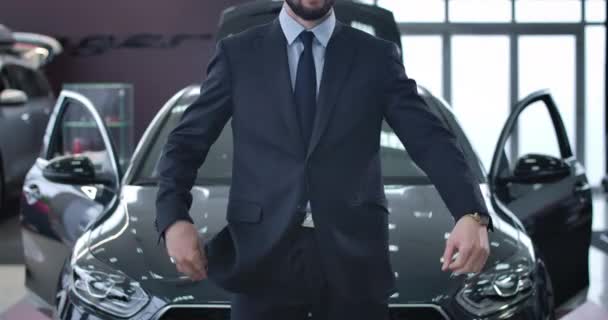 Молодой успешный кавказский мужчина в костюме стоит в салоне перед новой черной машиной. Бородатый бизнесмен покупает машину в выставочном зале. Автопромышленность, успех. Кино 4k proRes HQ . — стоковое видео