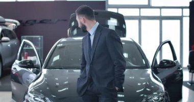 Yakışıklı genç adam yeni siyah arabaya bakıyor, kameraya dönüyor ve gülümsüyor. Beyaz, şık takım elbiseli bir işadamı otomobil galerisinde aracın önünde poz veriyor. Sinema 4k Prores Merkezi.