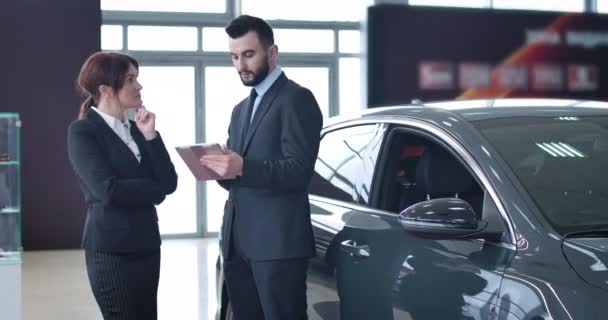 Young bearded Caucasian man consulting adult female client in car dealership. Confident woman talking with male trader in showroom. Automobile industry, selling, buying. Cinema 4k ProRes HQ. — Stock Video