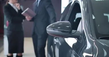 Arka planda iki bulanık beyaz adamın konuştuğu bir galeride duran siyah arabanın yakın çekimi. Tüccar ve müşteri galeride araç satın almayı tartışıyor. Sinema 4k Prores Merkezi.