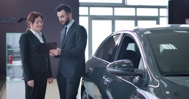 Позитивна успішна кавказька жінка підписує угоду про продаж і купівлю автомобілів. Молодий чоловік, який посміхається торговцю чоловіками, який обслуговує жінок у виставковому залі. Автомобільна промисловість. Фільм 4k Prores Hq. — стокове відео