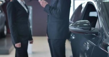 Tanımlanamayan beyaz bir erkek ve kadın arabanın anahtarlarını uzatıyor ve arka planda el sıkışıyor. Satıcılıkta ön planda duran siyah otomobillere odaklanmış. Sinema 4k Prores Merkezi.