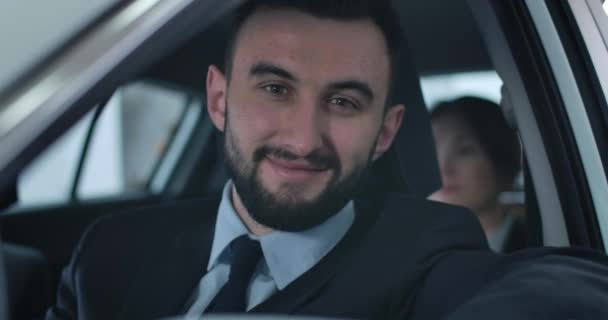 Gros plan sur le visage d'un jeune homme blanc beau qui regarde la caméra et sourit. Chauffeur professionnel assis sur le siège du conducteur dans la voiture avec passager à l'arrière-plan. Siège social Cinema 4k ProRes . — Video