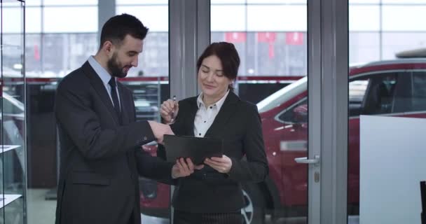 Representante de vendas caucasiano masculino dando contrato para assinar para empresária adulta comprando automóvel. Duas pessoas que fazem venda e compra acordo na concessionária de automóveis. Cinema 4k ProRes HQ . — Vídeo de Stock