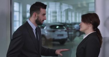 Ciddi bir beyaz erkek ve kadının araba galerisinde dikilip konuşmalarının yan görüntüsü. Erkek satıcı kadın müşteriye otomobil özelliklerini açıklıyor. Sinema 4k Prores Merkezi.