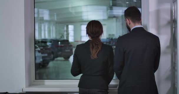 Πίσω όψη του ενήλικου Καυκάσου άνδρα και γυναίκα με κοστούμια κοιτάζοντας τα αυτοκίνητα στην αντιπροσωπεία και μιλώντας. Γυναίκα έμπορος συμβουλεύει αρσενικό πελάτη στην έκθεση. Αυτοκινητοβιομηχανία. Κινηματογράφος 4k Prores Hq. — Αρχείο Βίντεο