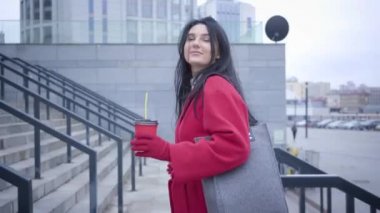Side view of positive brunette Caucasian girl walking up the stairs in city. Young beautiful woman in red coat strolling with coffee cup. Lifestyle, city life, happiness.