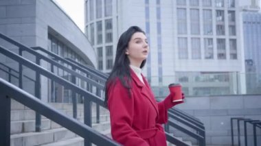 Camera following young pretty Caucasian woman walking down the stairs in city with coffee cup. Side view of elegant lady in red coat enjoying autumn day outdoors. Leisure, lifestyle, happiness.