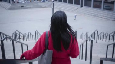 Back view of elegant young lady in red coat walking down the stairs. Confident Caucasian woman with black hair strolling in city in autumn day holding coffee cup. Lifestyle, break, leisure.