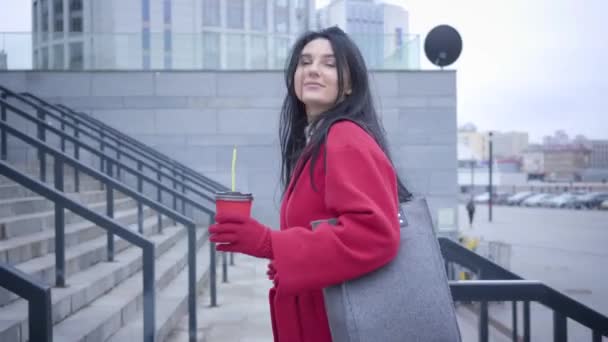 Side view of positive brunette Caucasian girl walking up the stairs in city. Young beautiful woman in red coat strolling with coffee cup. Lifestyle, city life, happiness. — 비디오