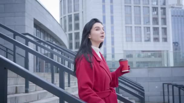 Câmera seguindo jovem mulher branca bonita descendo as escadas na cidade com xícara de café. Vista lateral da senhora elegante em casaco vermelho desfrutando de dia de outono ao ar livre. Lazer, estilo de vida, felicidade . — Vídeo de Stock