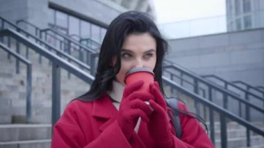 Portrait of young beautiful Caucasian woman with black hair drinking coffee in city. Smiling confident girl standing outdoors and looking around. City life, lifestyle, leisure.