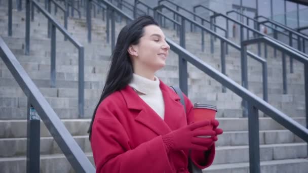 Profile portrait of charming Caucasian lady in red coat and gloves drinking coffee in city. Young cheerful woman standing on stairs outdoors and having break. Lifestyle, coffee break, leisure. — Stockvideo