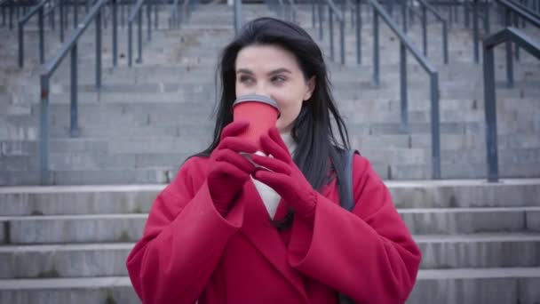 Portrait d'une jeune femme joyeuse buvant du café et étirant la tasse à la caméra. Jolie fille positive en manteau rouge et gants ayant une pause à l'extérieur. Loisirs, mode de vie, vie urbaine . — Video