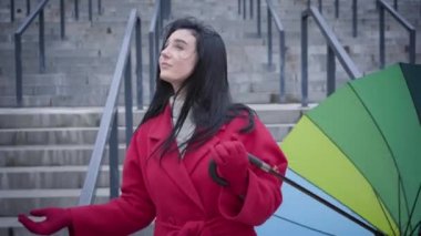 Pretty cheerful Caucasian woman folding umbrella and looking at camera. Joyful lady in red coat enjoying light rain outdoors in city. Happiness, lifestyle, leisure.