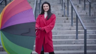 Portrait of cheerful Caucasian woman opening and spinning umbrella. Happy young lady in red coat smiling at camera holding multi-colored umbrella. Joy, lifestyle, fun.