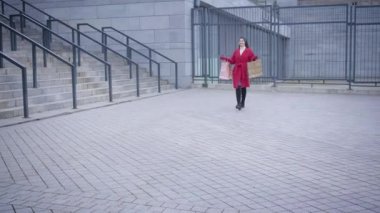 Young happy Caucasian woman in red coat and high black boots walking along the city square and leaving. Satisfied girl strolling with shopping bags. Shopping, fashion, lifestyle.