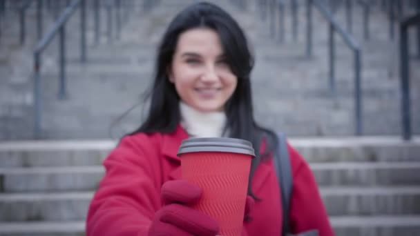 Невпізнавана розмита кавказька жінка розтягує чашку кави до камери і посміхається. Молода пані в червоному пальто і рукавички, які насолоджуються проривом по сходах у місті. Міське життя, спосіб життя. Зосереджений на чашці. — стокове відео