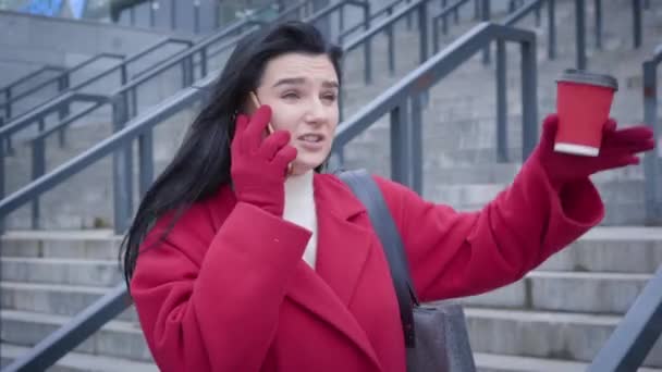 Camera following young Caucasian woman talking on the phone outdoors. Stylish girl in red coat and gloves waiting for meeting on city street. Beauty, lifestyle, leisure. — ストック動画